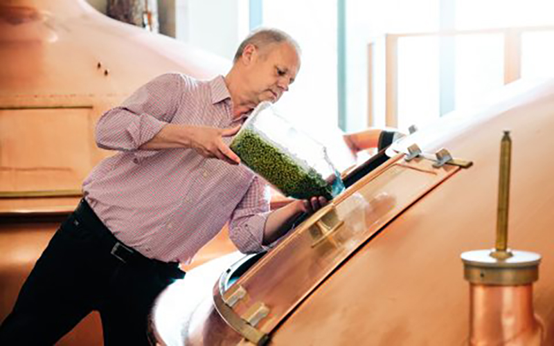 Bild von einem Bierbraumeister in der Hirschbrauerei, der gerade aus einem großen Glas grüne Zusätze in den Braukessel füllt