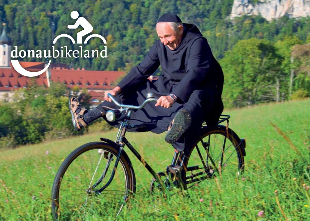 Donaubergland Postkartenmotiv mit fahrradfahrendem Mönch, der die Beine in die Luft hält und oberhalb eines Klosters in der Wiese fährt.