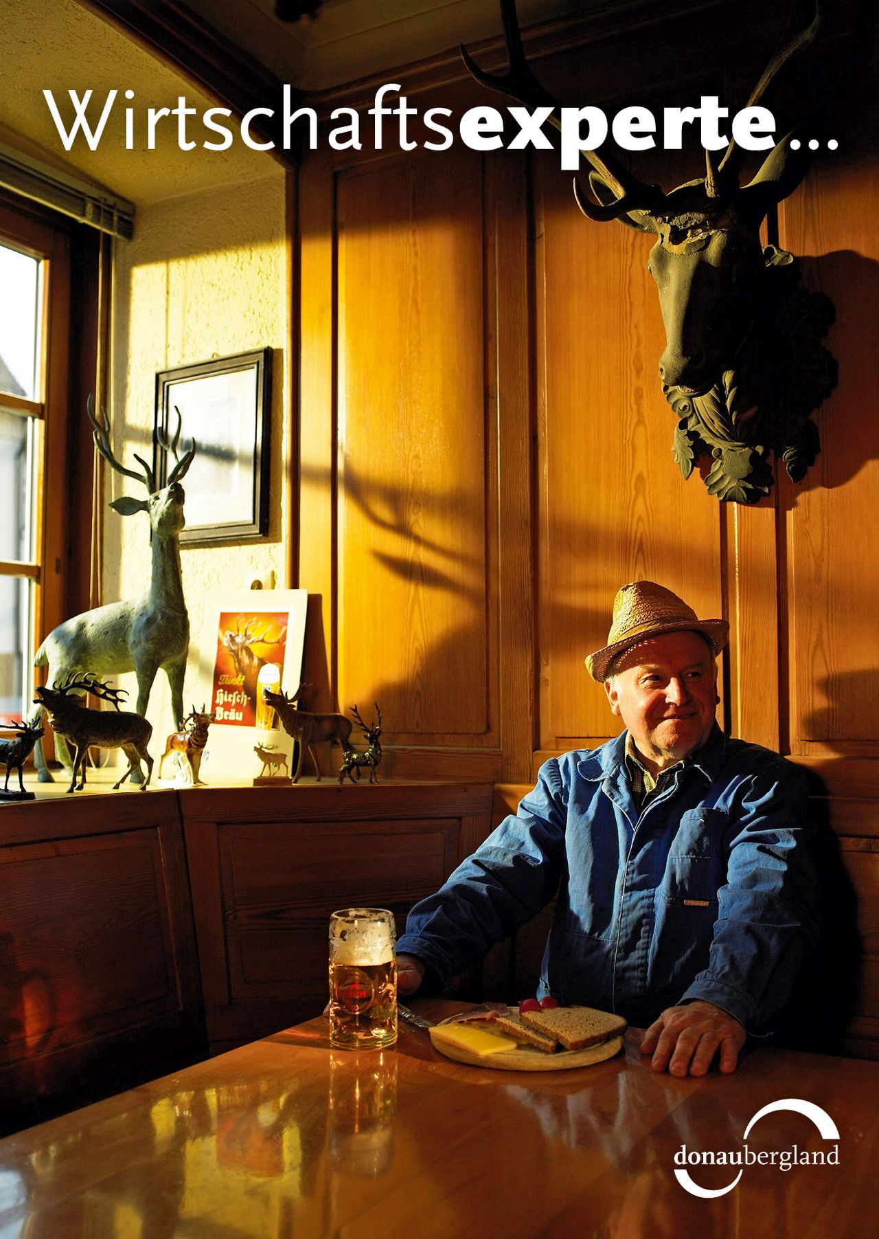 Donaubergland Postkartenmotiv mit Mann mit Hut in einer Wirtschaft mit Vesperbrett und Bier vor sich und über Ihm hängt ein Hirschgeweih und auf dem Fenstersims stehen ebenfalls Hirsche in verschiedenen Größen
