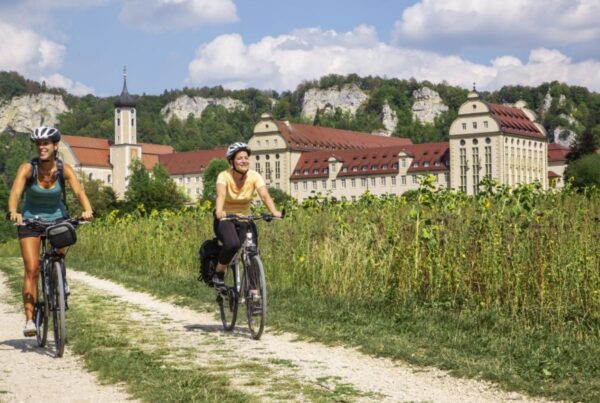 Bild von zwei Radfahrerinnen vor dem Kloster Beuron mit Felsen im Hintergrund auf einem Radweg mit Blumenwiese am Wegrand