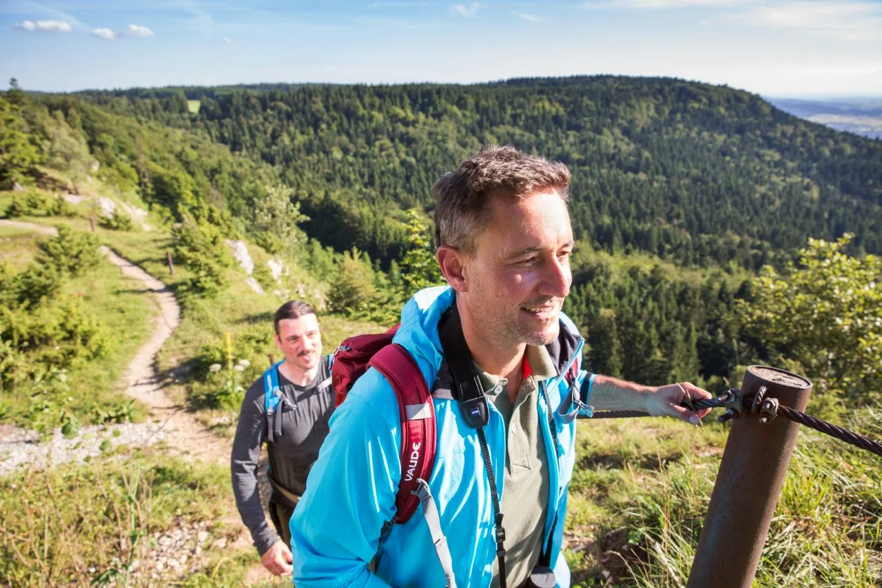 Bild von zwei Wanderern am Albtrauf, die hintereinander eine Anhöhe hinaufgehen hinter Ihnen liegt ein schmaler Wanderweg und ringsherum sind Bäume zu sehen