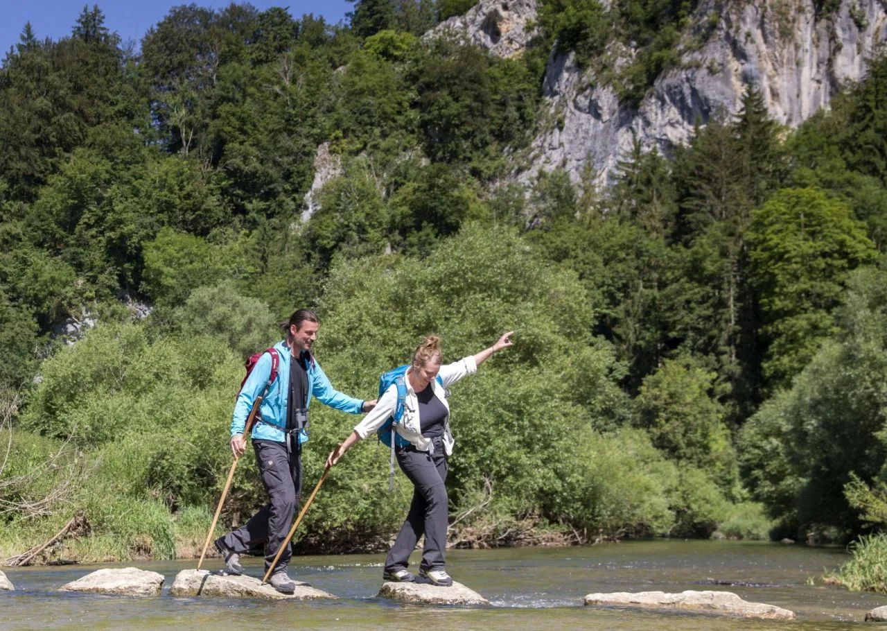 Bild von zwei Wanderern auf der Donaufelsentour, die auf Felsen die Donau überqueren. Beide haben einen Wanderstock in der Hand