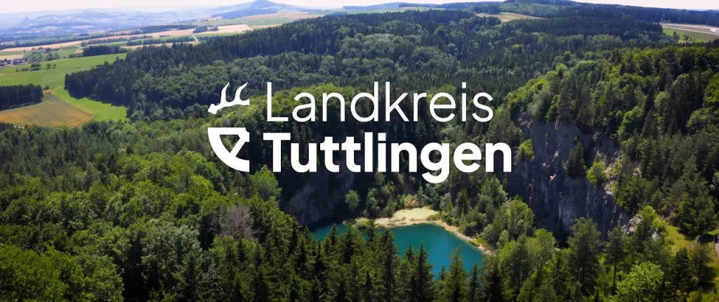 Videovorschaubild mit Blick von oben auf türkisfarbenen See, Bäumen, Feldern und Bergen im Hintergrund