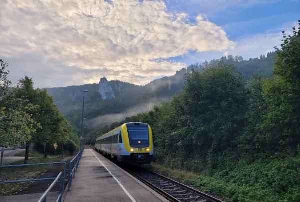 Bild eines einfahrenden Zugs in Beuron am Bahngleis. Im Hintergrund ist ein Felsen in der Höhe mit Kreuz zu erkennen