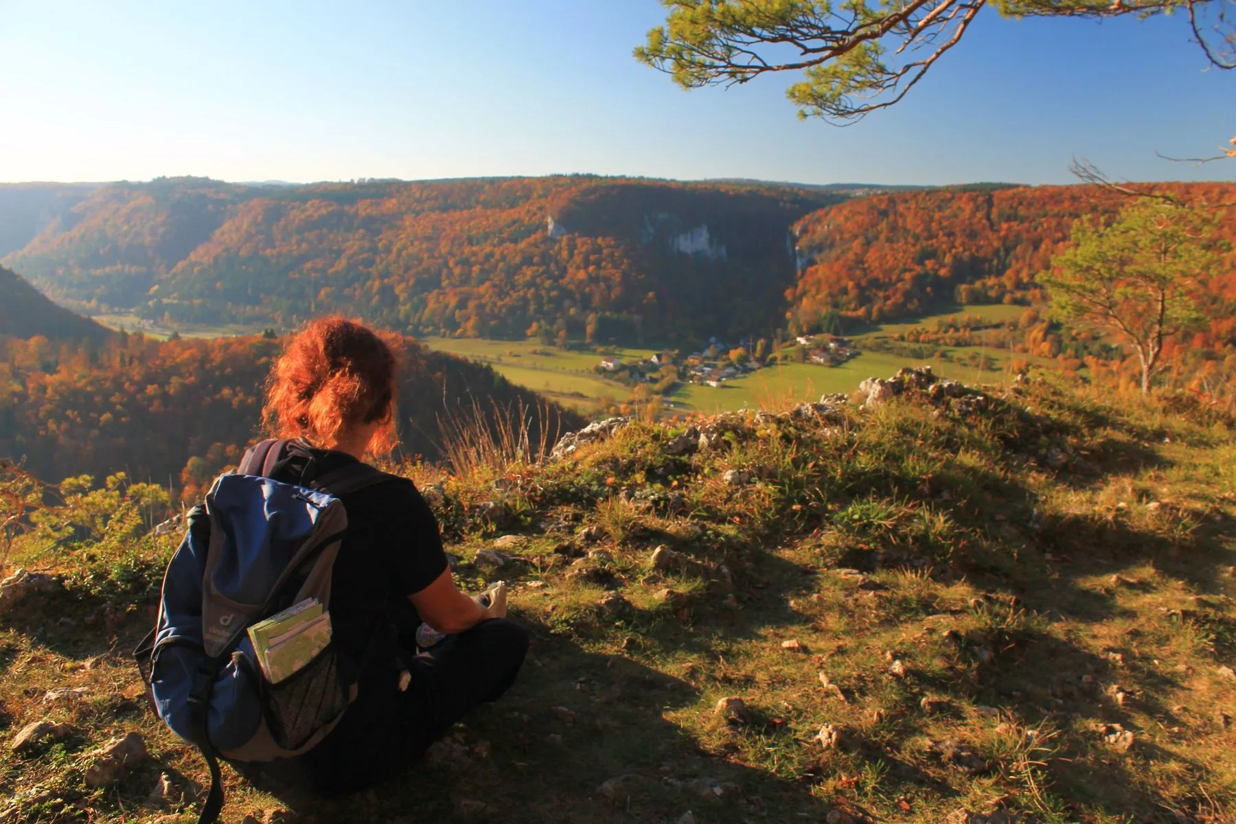 Eine Frau mit Rucksack sitzt auf felsigem Aussichtspunkt und schaut in eine herbstlich gefärbte Landschaft